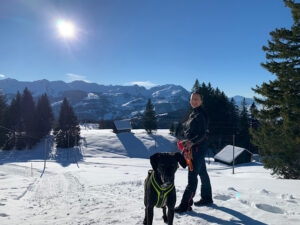 Hund Amor und Petra stehen im Schnee vor herrlichem Winterpanorama und den Schneebergen