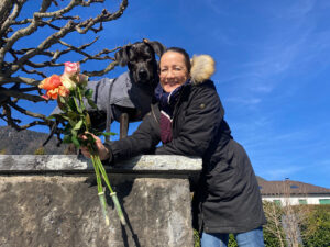 Petra lehnt an die Seeufersteinwand hält 3 Rosen in der Hand, der schwarze Hund Amor schaut erstaunt