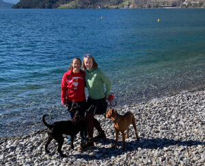 Petra steht mit Freundin Jana und der rotenfelligen Weslahündin Joko und dem schwarzen Kurzhaarmischlingshund Amor am steinigen Seeufer