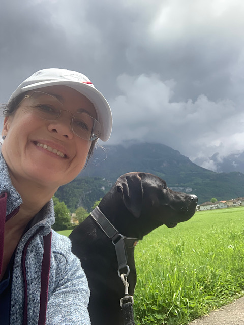 Petra mit Hund Amor beim Mittagsspaziergang, dunkle Regenwolken hängen am Berg, sie beide sind von Sonne beleuchtet