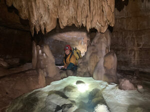 Petra sitzt in einer Höhle und wird beleuchtet, sie lächelt und sieht in die Kamera. Über ihr sind Tropfsteine, vor ihr ein Sintersee.