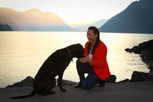 Petra bei Sonnenuntergang am See mit Hund Amor. Der Hund sitzt am Quaiufer und Petra kniet und hält Amor einen Futterbeutel unter die Schnauze.
