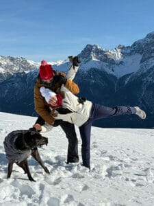 Petra mit Partner und Hund Amor blödeln und turnen im Schnee. Verschneite Bergkette im Hintergrund mit blauem Himmel