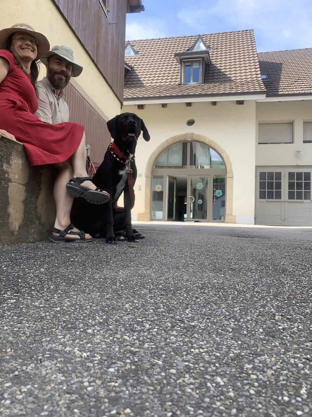 Zwei Menschen mit schwarzem Hund sitzen vor dem Eingang eines Hauses. Im Hintergrund ist der schöne Eingang einer Gesundheitspraxis