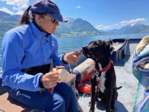 Petra sitzt mit schwarzem Hund Amor im Boot das auf dem Vierwaldstättersee fährt. Amor, der schwarze Hund liebt die leckere Hundewurst die er Rädchen um Rädchen gefüttert bekommt.