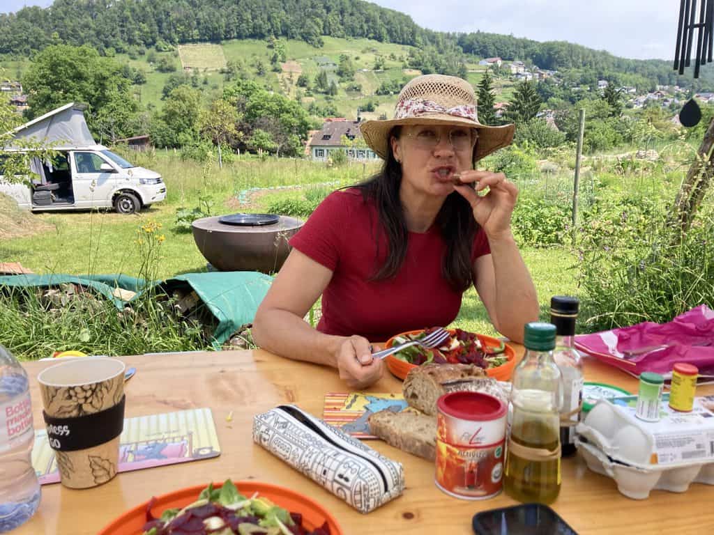 Frau im roten Kleid sitzt am Gartentisch im Naturkarten beim Mittagessen. Es gibt Salat.