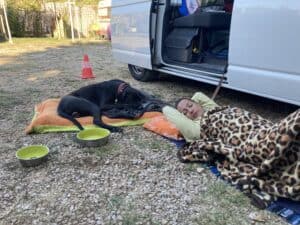 schwarzer Hund und Petra erholen sich liegend auf dem Campingplatz vor dem Bus am Boden liegend